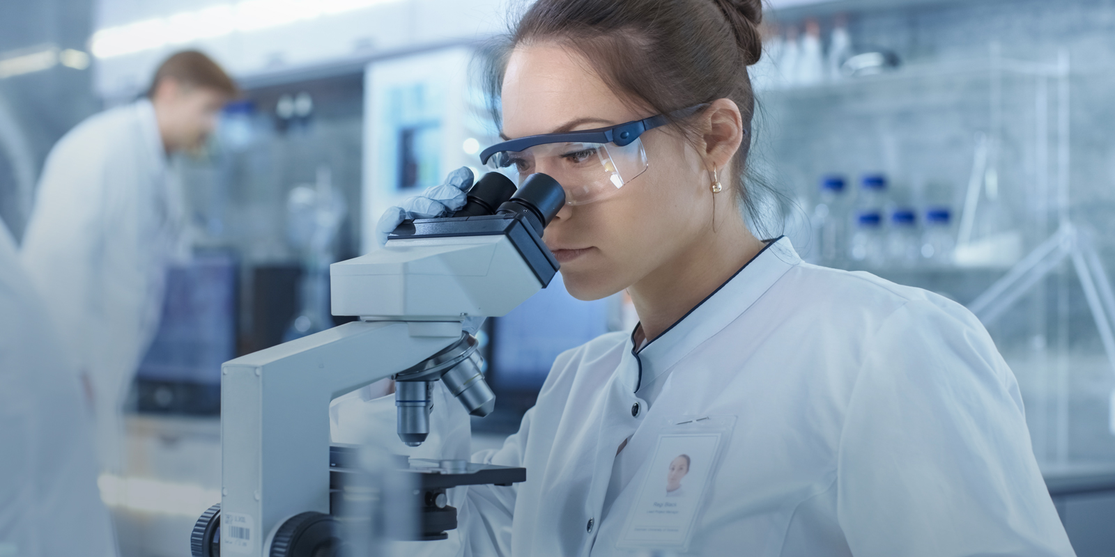 Person wearing safety glasses and lab coat looks through microscope